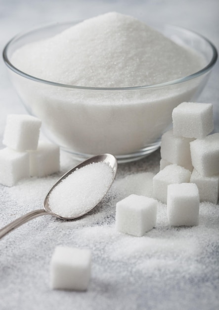 Glass bowl of natural white refined sugar with cubes and silver spoon on light background