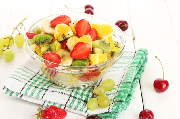 Glass bowl with fresh fruits salad and berries on white wooden table