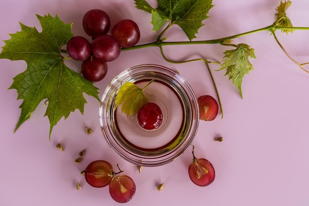 Glass bowl with organic grape seed oil ripe red grape berries\
top view lilac background high content of vitamins