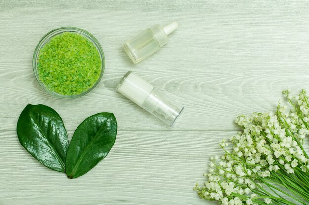 Glass bowl with sea salt, bottles with cream for face skin and aromatic oil, green leaves and bouquet of lilies of the valley on grey wooden background. Natural cosmetics for skin care