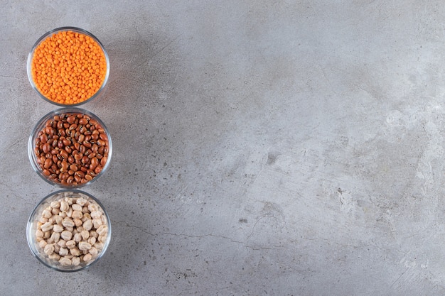 Photo glass bowls full of raw lentil, peas and beans on stone background.