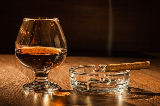 A glass of brandy and a cigar on the wooden table.