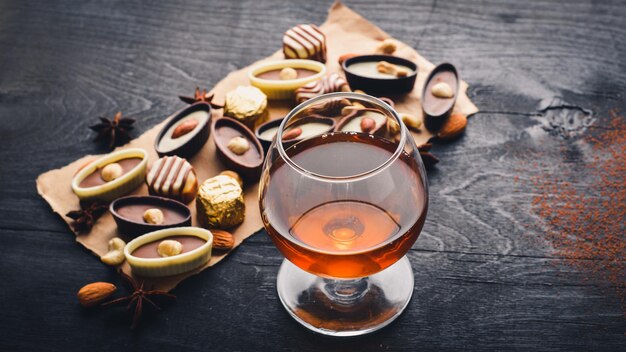 A glass of brandy Whiskey Assortment of chocolates Wooden on the black surface Top view