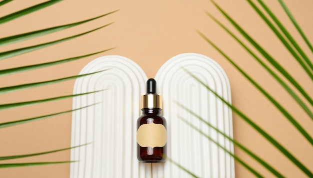 Glass brown bottle with pipette and paper blank label on light brown background, near palm leaf. Mockup skincare cosmetic product