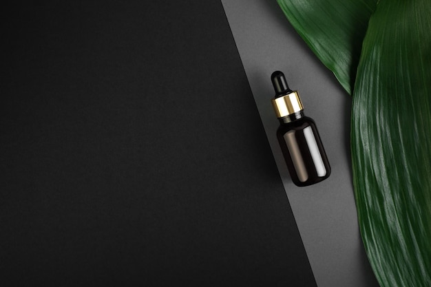 Glass brown bottle with pipette with oil on a grey black background with green tropical leaves