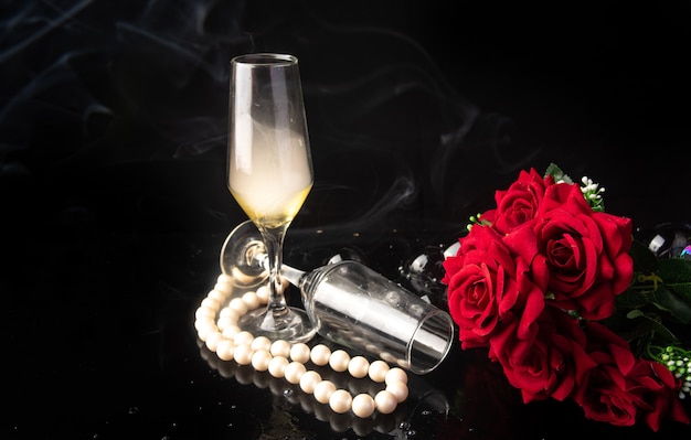 Glass of champagne, red roses, pearl necklace on black background, selective focus.