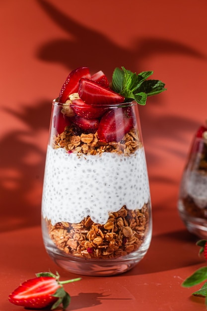 A glass of chia pudding with strawberries and granola on a red background.