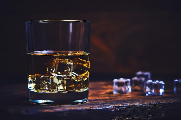 Glass chilled whiskey with ice cubes on wooden background in cellar.