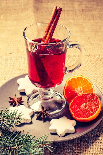クリスマスのグラスは、シナモン、スターアニス、クローブをプレートに、オレンジ、白のビスケット、天然のモミの木の枝を背景にしたグリューワインです。色調効果。