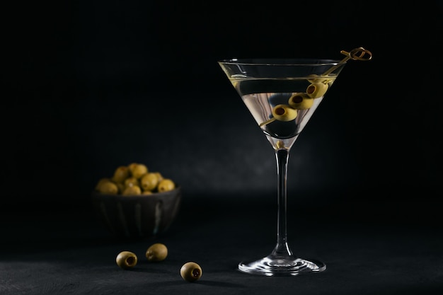 Glass of classic cocktail with olives on dark stone table against black surface. 