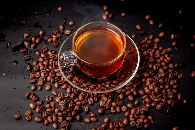 Glass of coffee on saucer and pile of roasted coffee bean scattered on black background, Hot dark coffee has smoke and aroma in cafe ,Drink for break, morning and coffee time ,beverage energy booster.