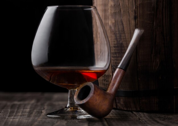 Glass of cognac brandy drink and vintage smoking pipe next to wooden barrel