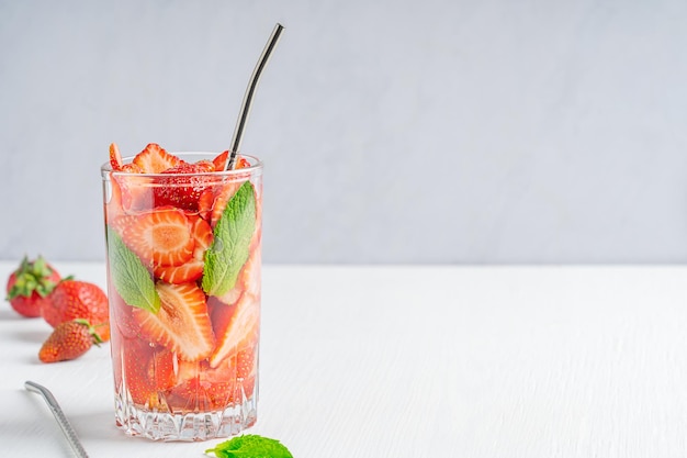 Bicchiere di acqua fredda detox infusa a base di fragola e foglia di menta servito con paglia sul tavolo bianco