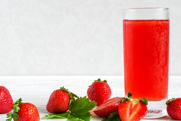 Glass of cold strawberry juice on white wooden table with ripe berries