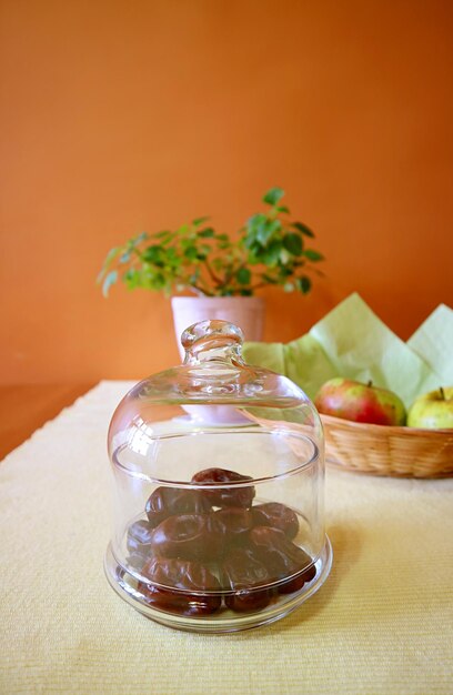 Glass compote bowl of dried dates on the table with blurry fruits and potted plant in background