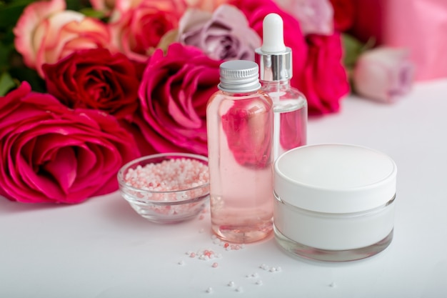 Glass cosmetic bottles, cream, serum, soap, oil on white table floral background. Flower red pink roses natural organic beauty product. Spa, skin care, bath body treatment. Set of cosmetics with rose.