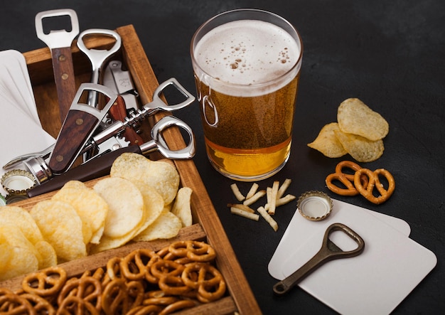 Photo glass of craft lager beer and opener with box of snacks on black kitchen table background pretzel and crisps and salty potato sticks in vintage wooden box with openers and beer mats