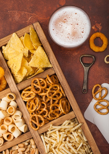 Glass of craft lager beer and opener with box of snacks on brown kitchen table. Pretzel,salty potato sticks, peanuts, onion rings with nachos in vintage box with openers and beer mats.