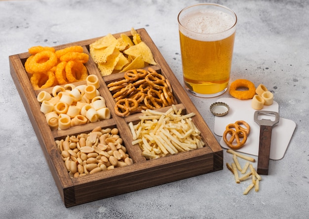 Photo glass of craft lager beer and opener with box of snacks on light kitchen table. pretzel,salty potato sticks, peanuts, onion rings with nachos in vintage box with openers and beer mats.