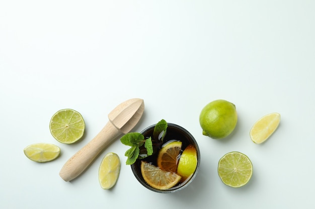 Glass of Cuba Libre, limes and wooden juicer on white surface, top view