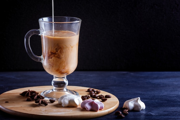 Glass cup of coffee with cream poured over and meringues on a wooden board on black