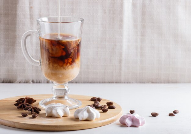 Glass cup of coffee with cream poured over and meringues on a wooden board on a white table.