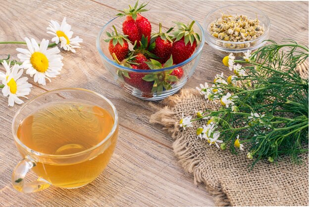 Glass cup of green tea, chamomile flowers, glass bowls with dry flowers of matricaria chamomilla and fresh strawberries on the wooden boards.