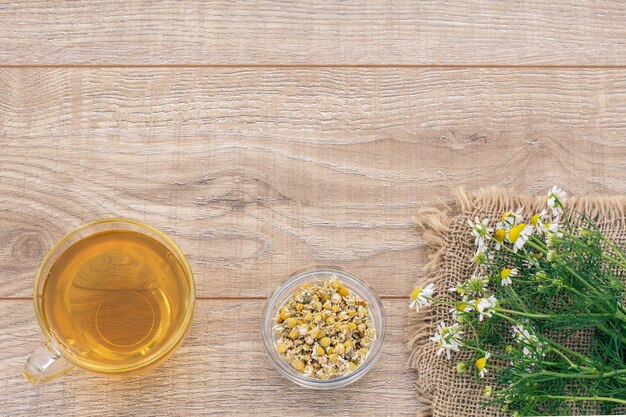 Glass cup of green tea, a little glass bowl with dry flowers of matricaria chamomilla and fresh chamomile flowers on the wooden background. Top view.