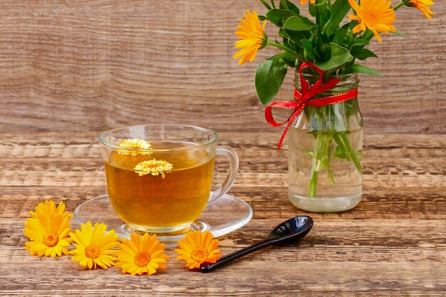 Photo glass cup of green tea with flowers of calendula and fresh bouquet of orange calendula in vase. wooden boards background. health-giving drink.