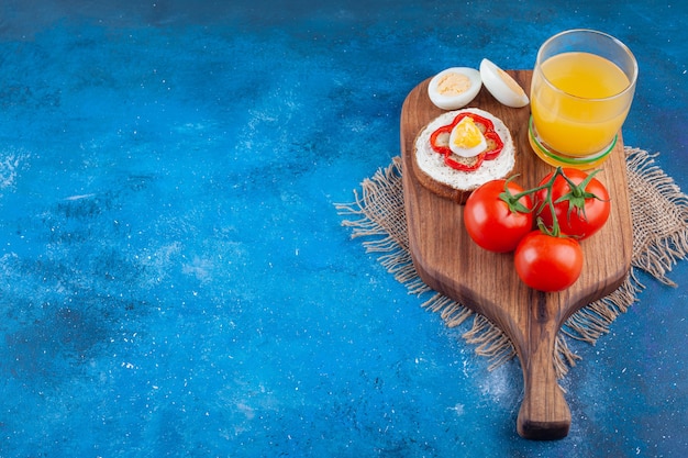 A glass cup of juice with sandwich and two red tomatoes on a wooden piece.