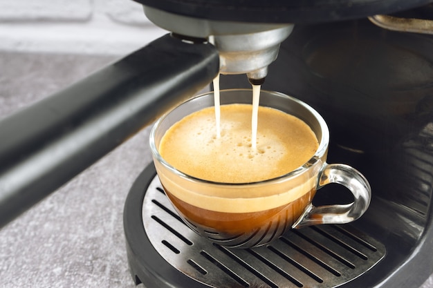 Glass cup standing on the grating of black coffee machine and coffee pouring into it. Process of preparing coffee.