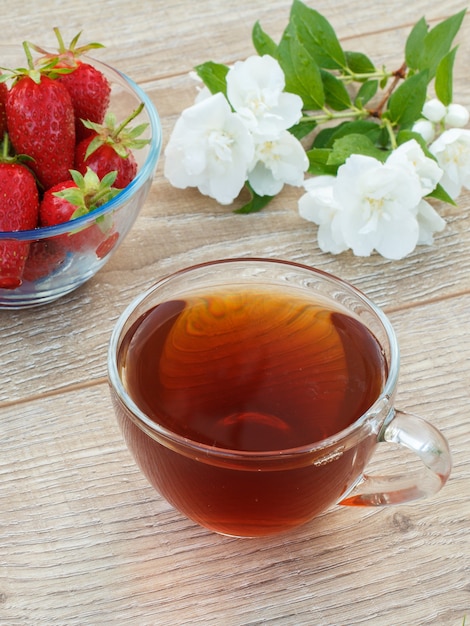 Glass cup of tea, bowl with fresh strawberries and white jasmine flowers on wooden background. Top view.