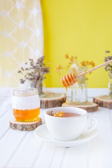 Tazza di tè di vetro con tiglio in erbe organiche naturali e un barattolo di miele su un tavolo di legno bianco