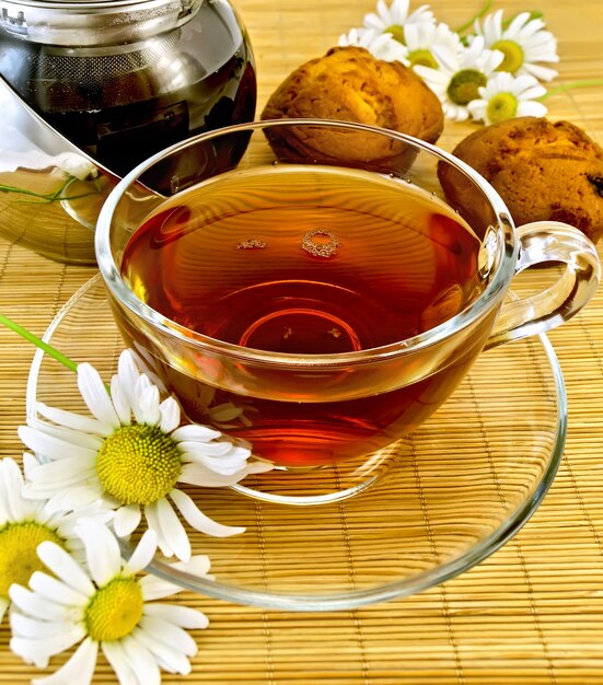 Glass cup with herbal tea, kettle, two cupcakes, camomile on a bamboo napkin