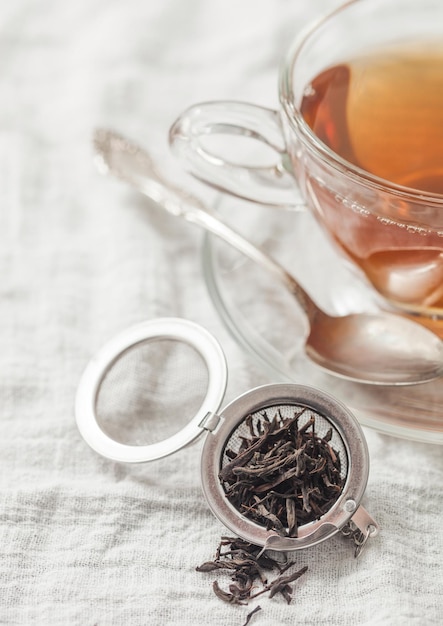 Glass cup with hot black tea and infuser with loose tea on\
white towel