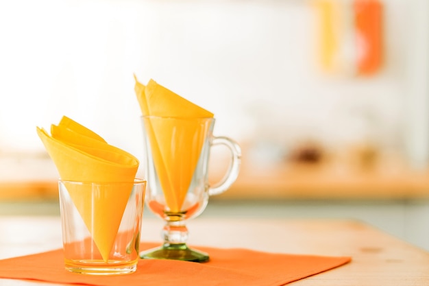 Glass cups decorated with yellow napkins stand on the table on orange napkins. Sunlight in the room