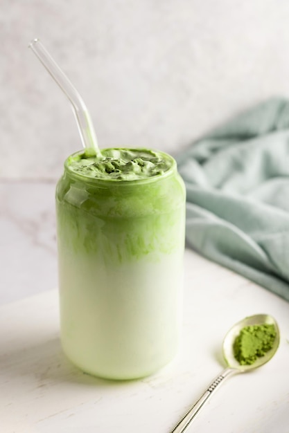 Glass of dalgona matcha latte and spoon of green tea powder on white background.