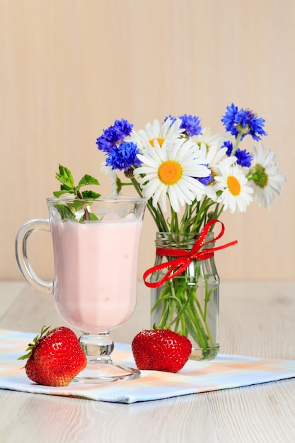 Glass of delicious yogurt with mint and fresh strawberries, chamomile and cornflowers in vase on a wooden table with a napkin