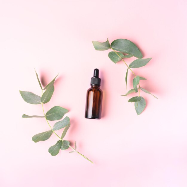 Glass dropper bottle and two branches of eucalyptus on pink background