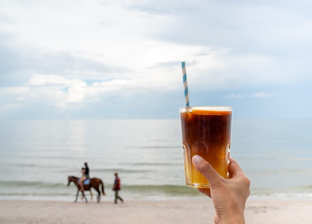 A glass of fresh iced orange americano decorated with sliced orange holding by hand on the beach background seascape view Cold black coffee with orange juice mixing mocktail drink menu