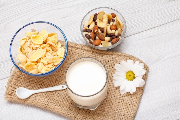 Glass of fresh milk, bowls with muesli and cornflakes, chamomile flower on sackcloth bag. Top view