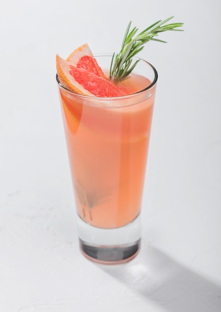 Glass of fresh pressed ruby red grapefruit juice with alcohol on white table background with rosemary and fruit slice.