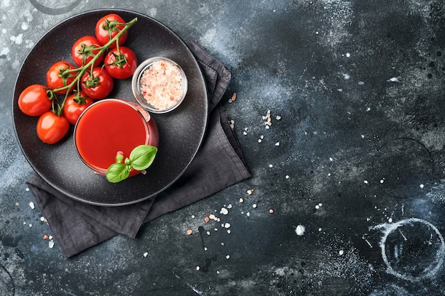 Glass of fresh tomato juice, salt, basil and tomatoes on black plate on old black background. Top view with copy space.