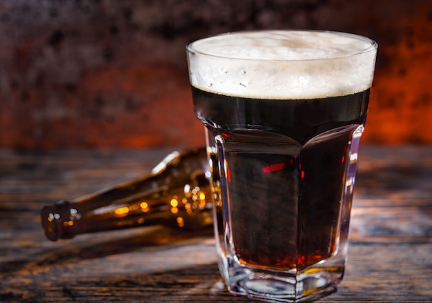 Glass of freshly poured dark beer near empty bottle on dark wooden desk. Food and beverages concept