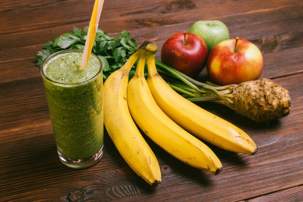 Glass of green smoothie with yellow straw on a wooden table next to bananas, apples and celery