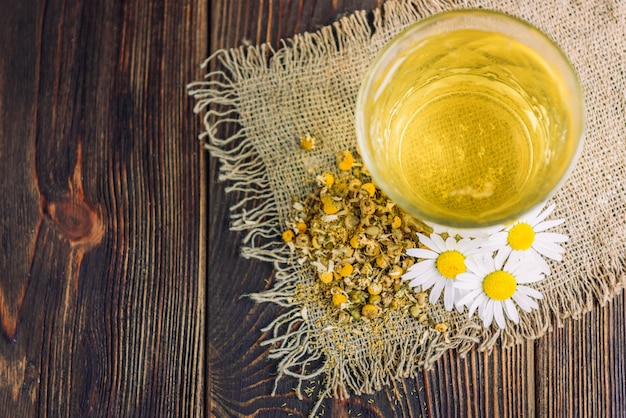 Glass of herbal tea with chamomile flowers on dark wooden background.