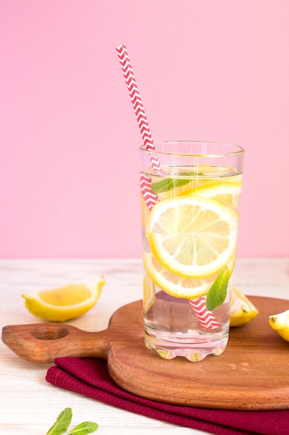 Glass of homemade lemonade with lemons, mint and red paper straw on pink background. Summer refreshing beverage.