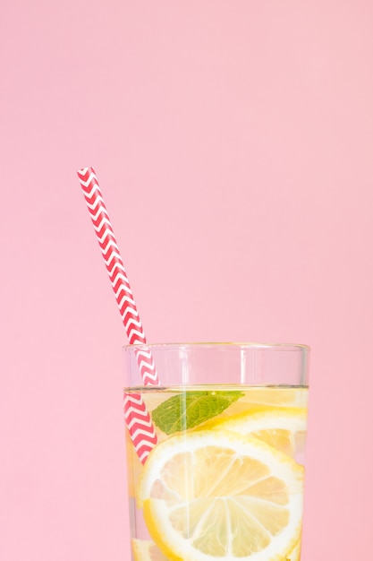 Glass of homemade lemonade with lemons, mint and red paper straw on pink background. Summer refreshing beverage.