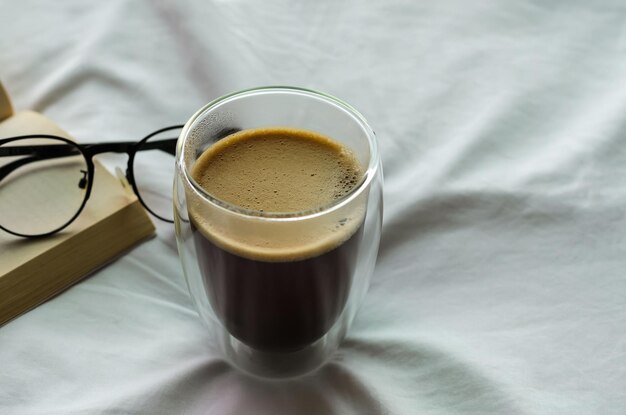 Photo a glass of hot coffee with book and spectacles on bed in morning stay home and relaxing concept