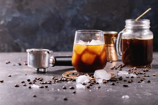 Glass of ice coffee with ice cubes served with cream, copper cezve and coffee beans over dark surface